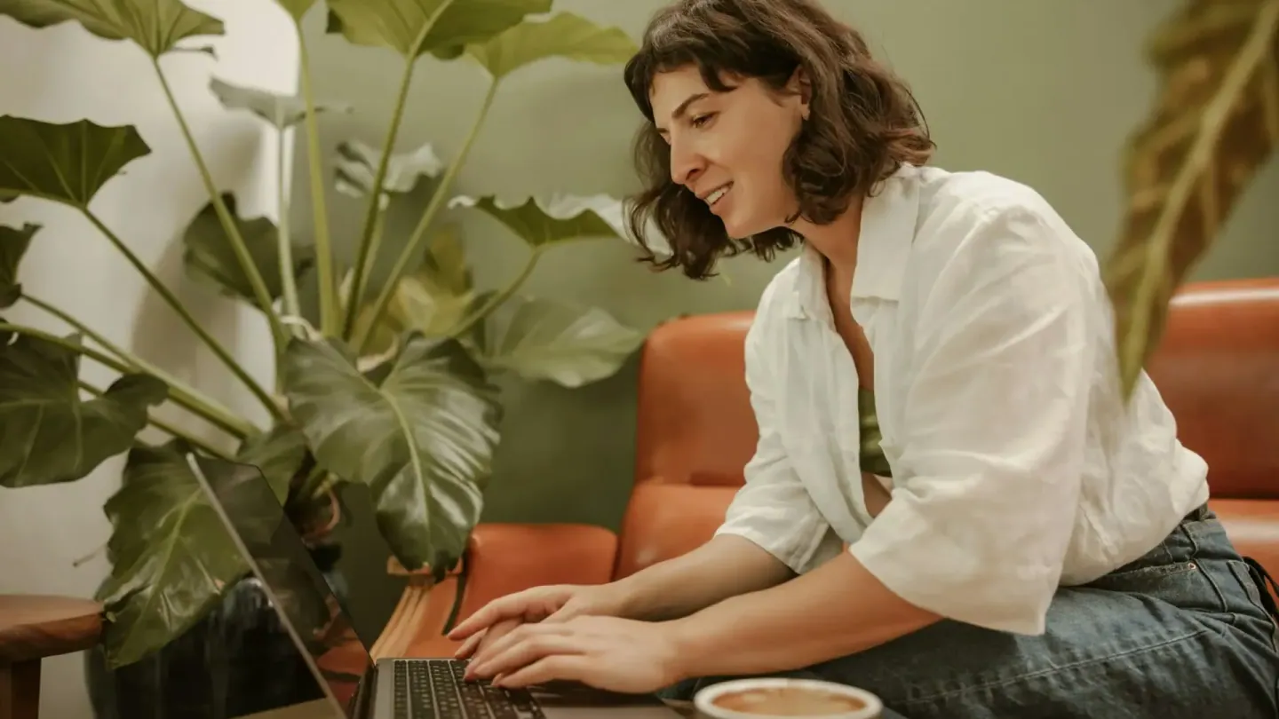 Een vrouw met halflang donker haar in een wit linnen overhemd op een cognackleurige bank tussen grote kamerplanten, voorovergebogen typend op een laptop op de salontafel, een kop koffie ernaast.