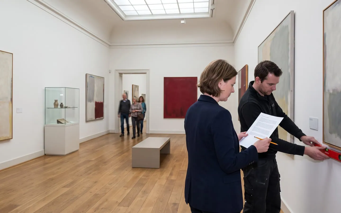 Een museumzaal, de conservator met zaalteksten naast een technicus met een waterpas, bezoekers verderop.