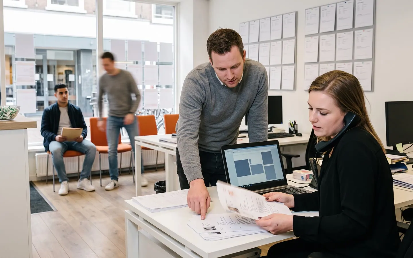 De front office van een uitzendbureau: een recruiter met de telefoon aan haar schouder en een stapel cv's, kandidaten op de stoelen.