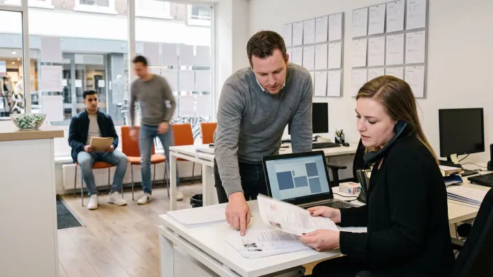 De front office van een uitzendbureau: een recruiter met de telefoon aan haar schouder en een stapel cv's, kandidaten op de stoelen.